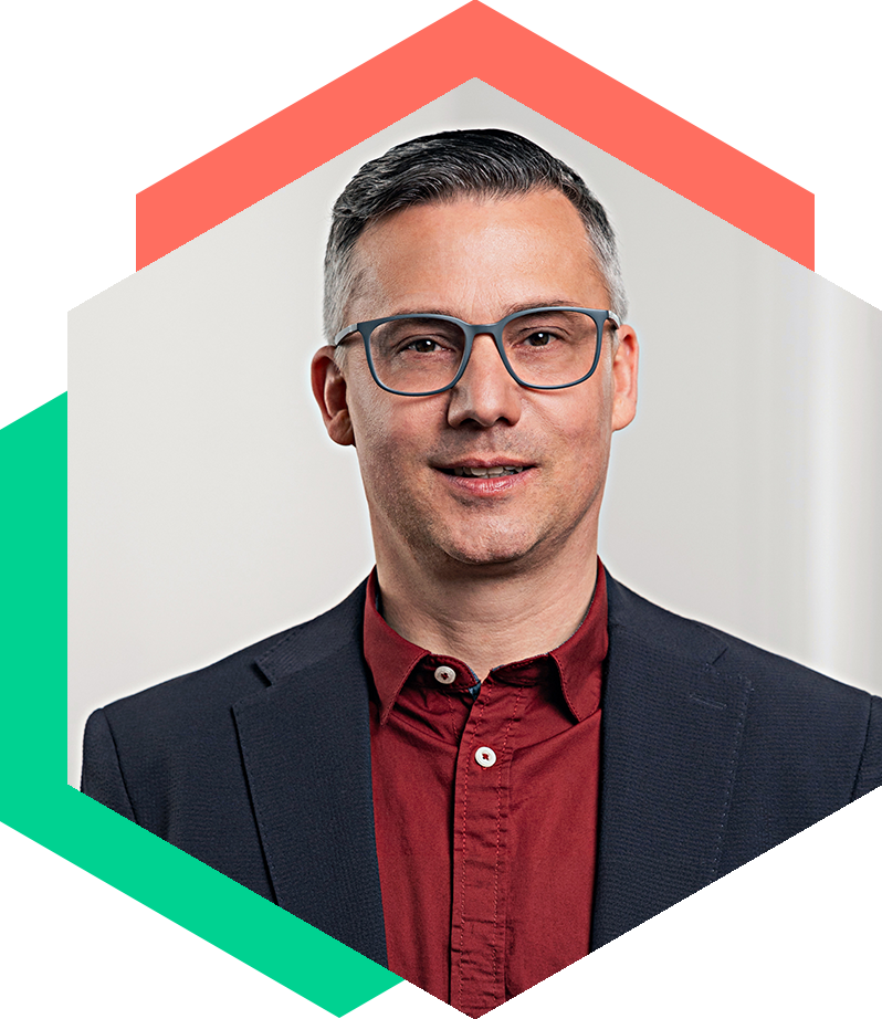 Porträt von Björn Fröbe mit Brille, dunklem Sakko und rotem Hemd, eingefasst in das hexagonale Design-Element – Senior Berater für strategische Kommunikation und Krisenmanagement bei WeichertMehner