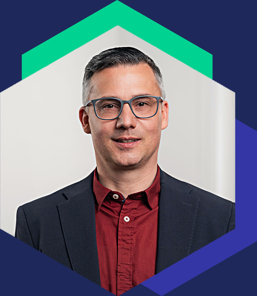 Porträt von Björn Fröbe mit Brille, dunklem Sakko und rotem Hemd, eingefasst in das hexagonale Design-Element – Senior Berater für strategische Kommunikation und Krisenmanagement bei WeichertMehner