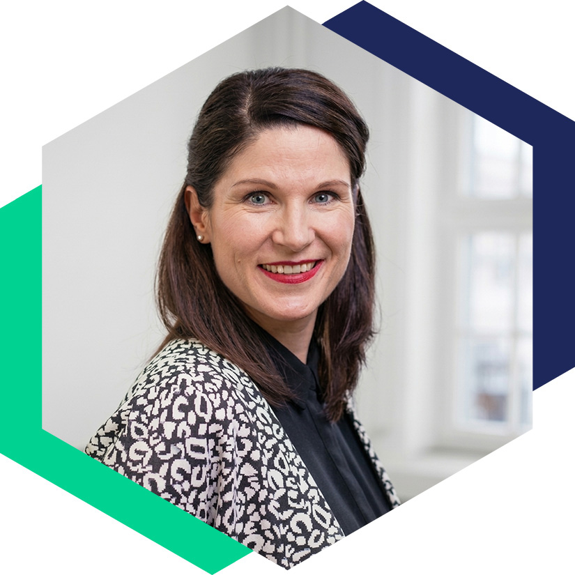 Porträt von Susann Smith in gemusterter Jacke vor hellem Hintergrund, grafisch eingerahmt in einem blauen Hexagon-Design, Senior Beraterin für strategische Kommunikation und Krisenmanagement bei WeichertMehner