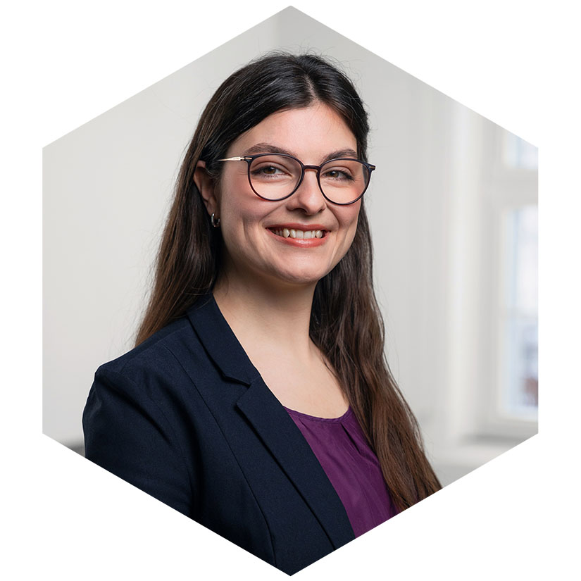 Porträt von Adriane Klaus mit Brille, dunklem Blazer und lila Oberteil, grafisch eingefasst in einem modernen Hexagon-Design, Beraterin für PR und strategische Kommunikation bei WeichertMehner