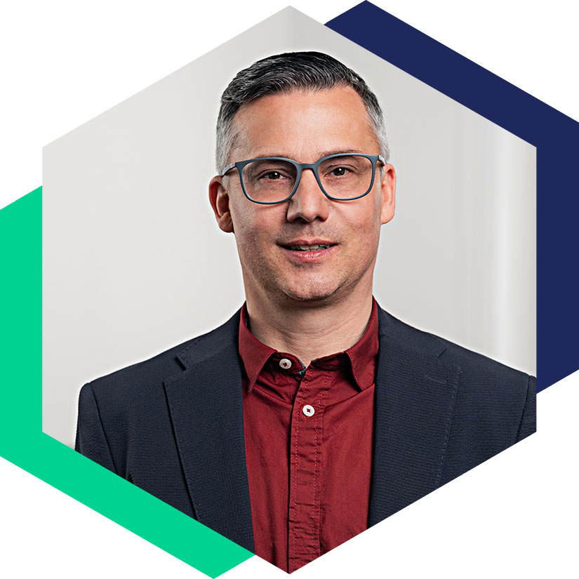 Porträt von Björn Fröbe mit Brille, dunklem Sakko und rotem Hemd, eingefasst in das hexagonale Design-Element – Senior Berater für strategische Kommunikation und Krisenmanagement bei WeichertMehner