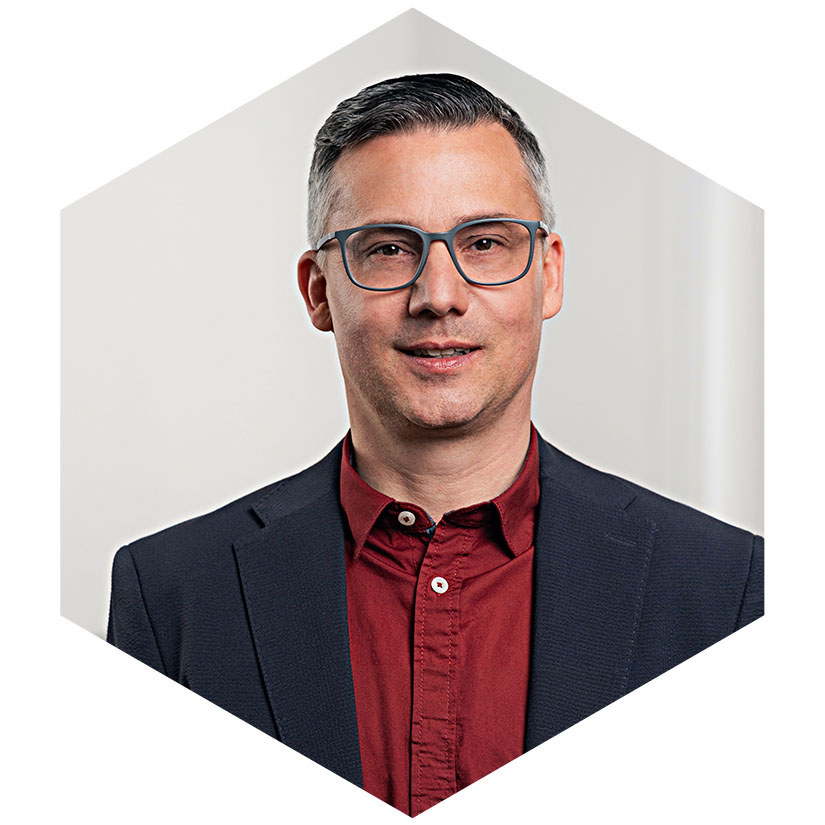 Porträt von Björn Fröbe mit Brille, dunklem Sakko und rotem Hemd, eingefasst in das hexagonale Design-Element – Senior Berater für strategische Kommunikation und Krisenmanagement bei WeichertMehner