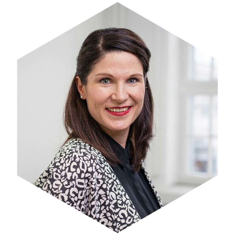 Porträt von Susann Smith in gemusterter Jacke vor hellem Hintergrund, grafisch eingerahmt in einem blauen Hexagon-Design, Senior Beraterin für strategische Kommunikation und Krisenmanagement bei WeichertMehner